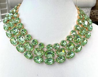 Può includere: Collana a cascata con tre file di cristalli verde chiaro, sfaccettati, ovali e rettangolari, incastonati in metallo dorato. La collana è esposta su un busto di manichino bianco.