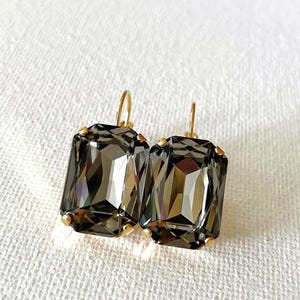 May include: A pair of rectangular, smoky gray crystal earrings with gold-colored hooks and settings. The faceted crystals reflect light, creating a sparkling effect. The earrings are displayed on a white, textured surface.