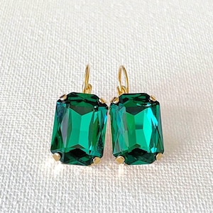 May include: A pair of emerald green rectangular earrings with gold-colored settings and ear wires. The earrings have a faceted design, reflecting light. The background is a white, textured fabric.