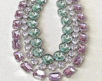 May include: A multi-strand necklace featuring rows of faceted crystals in shades of light purple, mint green, and clear white. The stones are set in silver-toned metal settings, arranged in a cascading design that creates a layered appearance.