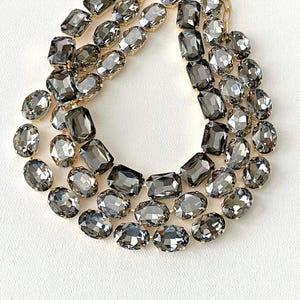 May include: A three-strand necklace featuring large, faceted gray gemstones in oval and rectangular shapes. The stones are set on a delicate gold-tone chain, creating a glamorous and eye-catching accessory. The necklace is displayed on a white surface.