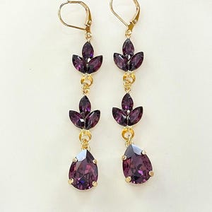 May include: A pair of long, dangling earrings with a gold setting and purple crystal accents. The earrings feature three teardrop-shaped crystals and a larger pear-shaped crystal at the bottom.