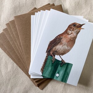 May include: A set of blank greeting cards with brown envelopes. Each card features a watercolor illustration of a brown wren perched on a green fence post, singing.