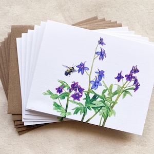 May include: A set of blank greeting cards featuring a watercolor illustration of purple and blue flowers with a bee flying nearby. The cards are packaged with brown envelopes.