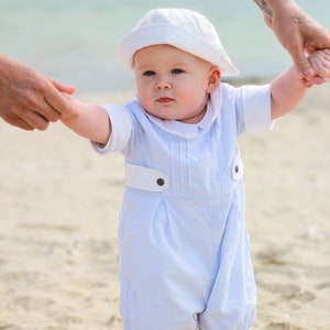 May include: A baby wearing a white sun hat and a light blue romper is taking their first steps on a sandy beach.