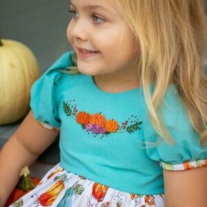 以下が含まれることがあります： かぼちゃと葉っぱが刺繍されたターコイズブルーのドレスを着た少女。ドレスは、かぼちゃ柄の白いスカートです。少女はブロンドの長い髪をしていて、笑顔です。