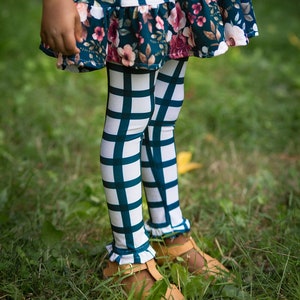 May include: Teal and white checkered leggings with a ruffled hem. The leggings are worn by a child and are shown against a green grassy background.