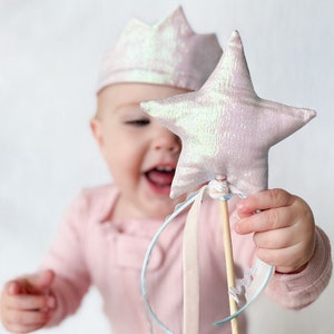 May include: A pink and white star-shaped wand with a wooden stick and blue and white ribbons. The wand is held by a child wearing a pink crown.