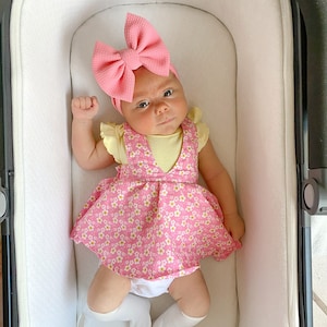May include: A baby wearing a pink bow headband, a yellow sleeveless shirt, and a pink floral dress with white flowers. The baby is lying in a white stroller.