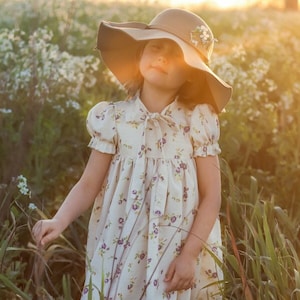 Könnte beinhalten: Ein junges Mädchen in einem weißen Kleid mit Blumenmuster und einem braunen Hut mit breiter Krempe steht in einem Feld mit hohem Gras.