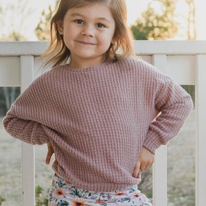 May include: A young child wearing a mauve waffle-knit long-sleeve top and a floral print skirt. The skirt features a pattern of orange, pink, and yellow flowers. The child is smiling and standing outside.