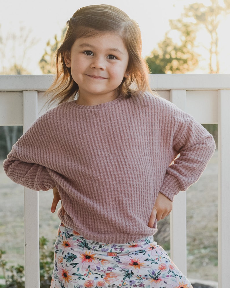 May include: A young child wearing a mauve waffle-knit long-sleeve top and a floral print skirt. The skirt features a pattern of orange, pink, and yellow flowers. The child is smiling and standing outside.