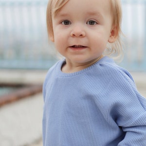 May include: A young child with blonde hair and light eyes is wearing a light blue waffle-knit long-sleeve shirt. The child is looking directly at the camera with a slight smile. The background is blurred, with a hint of a light blue structure.
