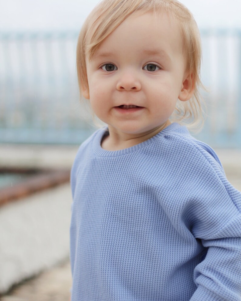 May include: A young child with blonde hair and light eyes is wearing a light blue waffle-knit long-sleeve shirt. The child is looking directly at the camera with a slight smile. The background is blurred, with a hint of a light blue structure.