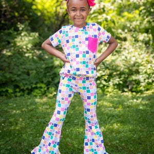 May include: A young person wearing a colorful, patterned outfit. The outfit includes a short-sleeved top with a pink pocket and flared pants. The pattern features squares with flowers in various colors. A pink bow is in their hair.