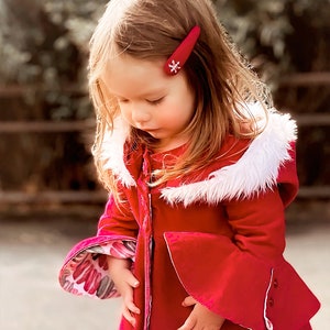 Pu&ograve; includere: Una bambina indossa un cappotto di velluto rosso con un bordo di pelliccia bianca intorno al cappuccio. Il cappotto ha una cerniera sul davanti e un fermaglio per capelli a forma di fiocco di neve nei capelli.
