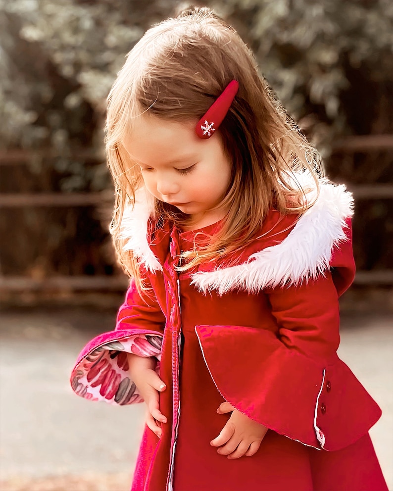 Pu&ograve; includere: Una bambina indossa un cappotto di velluto rosso con un bordo di pelliccia bianca intorno al cappuccio. Il cappotto ha una cerniera sul davanti e un fermaglio per capelli a forma di fiocco di neve nei capelli.