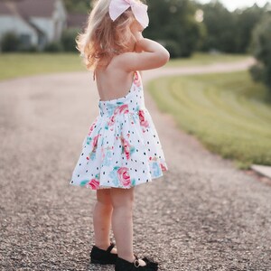 K&ouml;nnte beinhalten: Ein junges M&auml;dchen tr&auml;gt ein wei&szlig;es Kleid mit Blumen- und Tupfenmuster. Sie tr&auml;gt schwarze Schuhe und eine rosa Schleife im Haar.