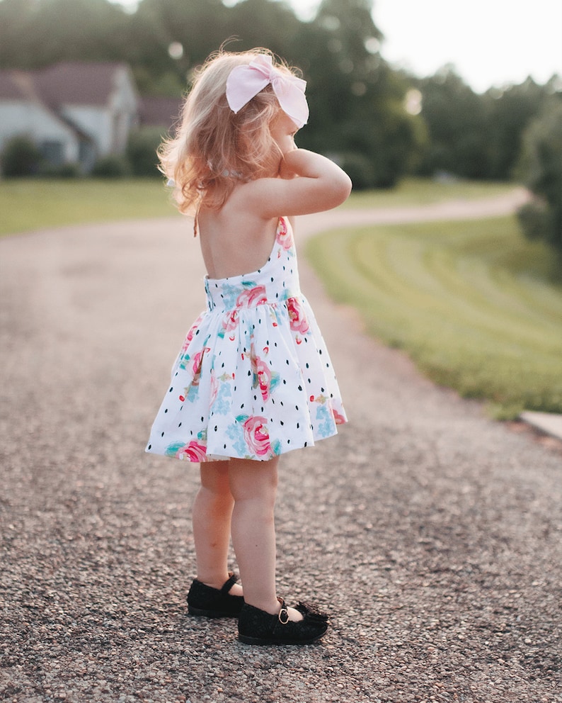K&ouml;nnte beinhalten: Ein junges M&auml;dchen tr&auml;gt ein wei&szlig;es Kleid mit Blumen- und Tupfenmuster. Sie tr&auml;gt schwarze Schuhe und eine rosa Schleife im Haar.