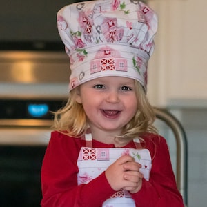 May include: A young child wearing a red long-sleeved shirt and a white and pink patterned apron with a matching chef's hat. The apron has a red and white checkered bottom and a pink heart design.