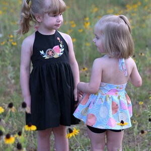 K&ouml;nnte beinhalten: Zwei junge M&auml;dchen in Sommerkleidern stehen auf einem Feld mit gelben Blumen. Das M&auml;dchen links tr&auml;gt ein schwarzes Halfterkleid mit einem Zitrusfruchtmuster. Das M&auml;dchen rechts tr&auml;gt ein hellblaues Kleid mit einem pinkfarbenen und gelben Zitrusfruchtmuster.