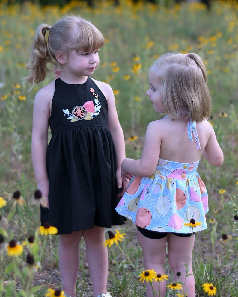 K&ouml;nnte beinhalten: Zwei junge M&auml;dchen in Sommerkleidern stehen auf einem Feld mit gelben Blumen. Das M&auml;dchen links tr&auml;gt ein schwarzes Halfterkleid mit einem Zitrusfruchtmuster. Das M&auml;dchen rechts tr&auml;gt ein hellblaues Kleid mit einem pinkfarbenen und gelben Zitrusfruchtmuster.