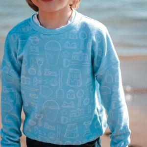 May include: Light blue sweatshirt with a white outline pattern of sandcastle building tools, buckets, and seashells. The long-sleeved top is a crew neck style. The sweatshirt is worn by a person.