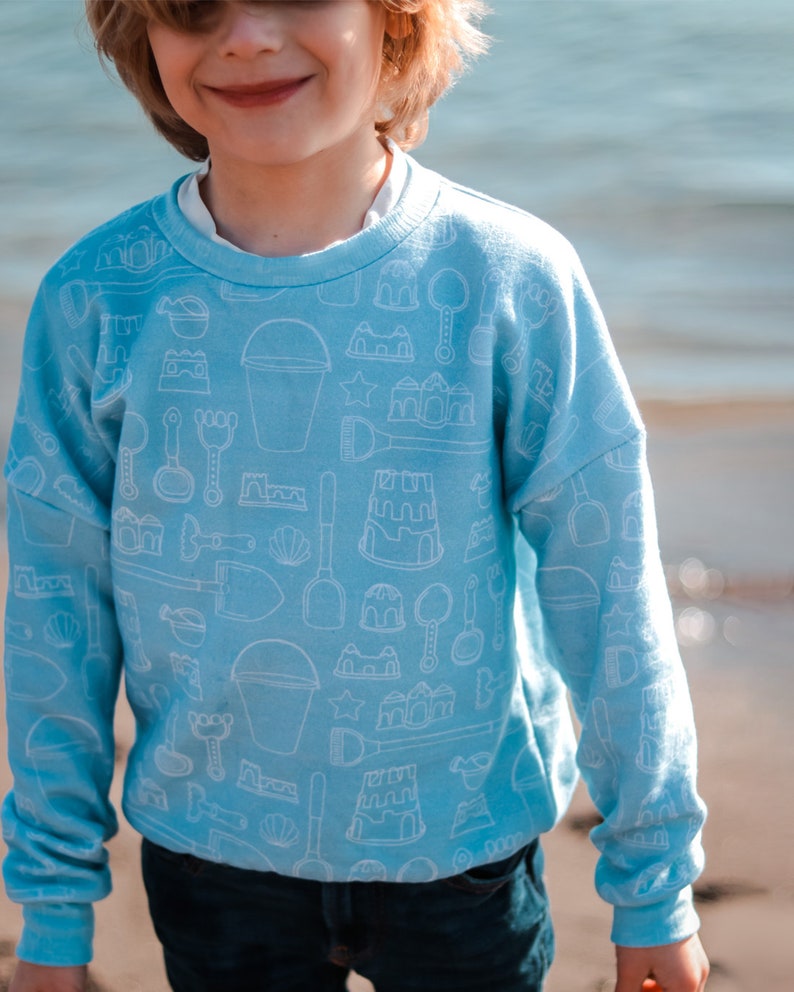 May include: Light blue sweatshirt with a white outline pattern of sandcastle building tools, buckets, and seashells. The long-sleeved top is a crew neck style. The sweatshirt is worn by a person.