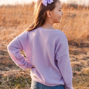 May include: A young person wearing a light purple waffle-knit sweater and blue jeans. A light purple bow with white polka dots is in their hair. The background is a field of dry grass.