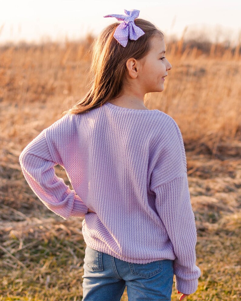 May include: A young person wearing a light purple waffle-knit sweater and blue jeans. A light purple bow with white polka dots is in their hair. The background is a field of dry grass.
