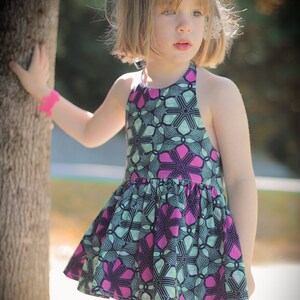 K&ouml;nnte beinhalten: Ein junges M&auml;dchen tr&auml;gt ein &auml;rmelloses Kleid mit einem pink- und gr&uuml;nfarbigen Blumenmuster. Das Kleid hat einen gerafften Rock und einen Halterneck.