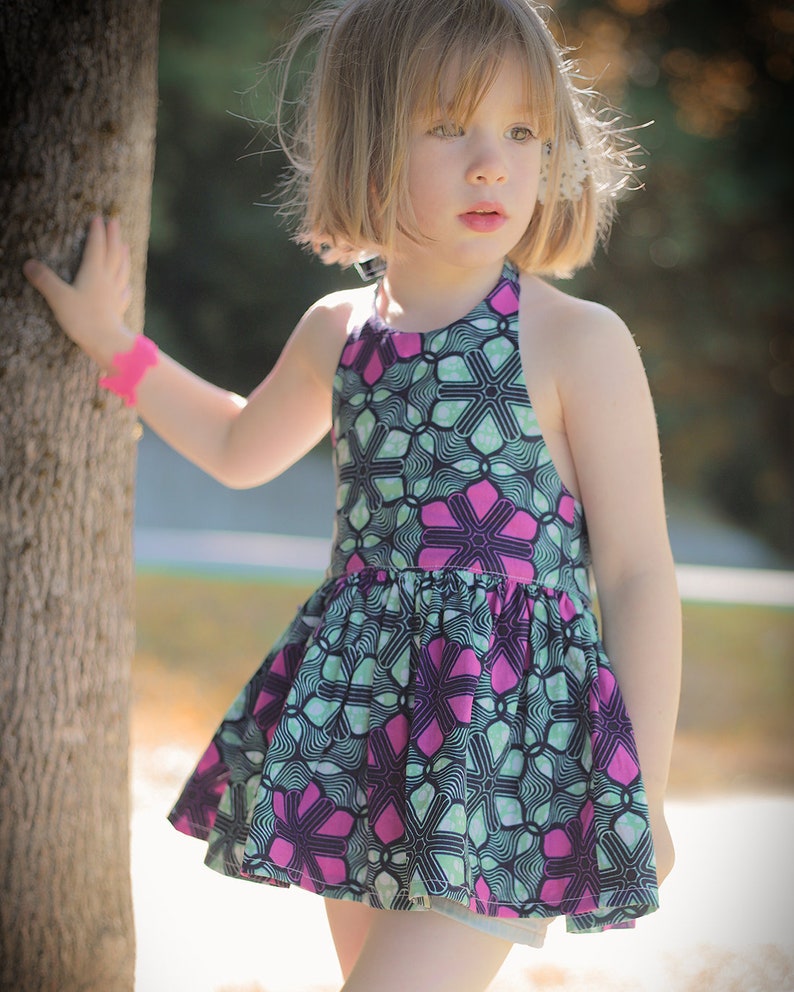 K&ouml;nnte beinhalten: Ein junges M&auml;dchen tr&auml;gt ein &auml;rmelloses Kleid mit einem pink- und gr&uuml;nfarbigen Blumenmuster. Das Kleid hat einen gerafften Rock und einen Halterneck.