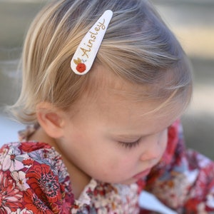 Pu&ograve; includere: Un fermaglio per capelli bianco con il nome "Ainsley" ricamato in filo dorato. Un piccolo fiore rosso con dettagli dorati &egrave; ricamato sul fermaglio.