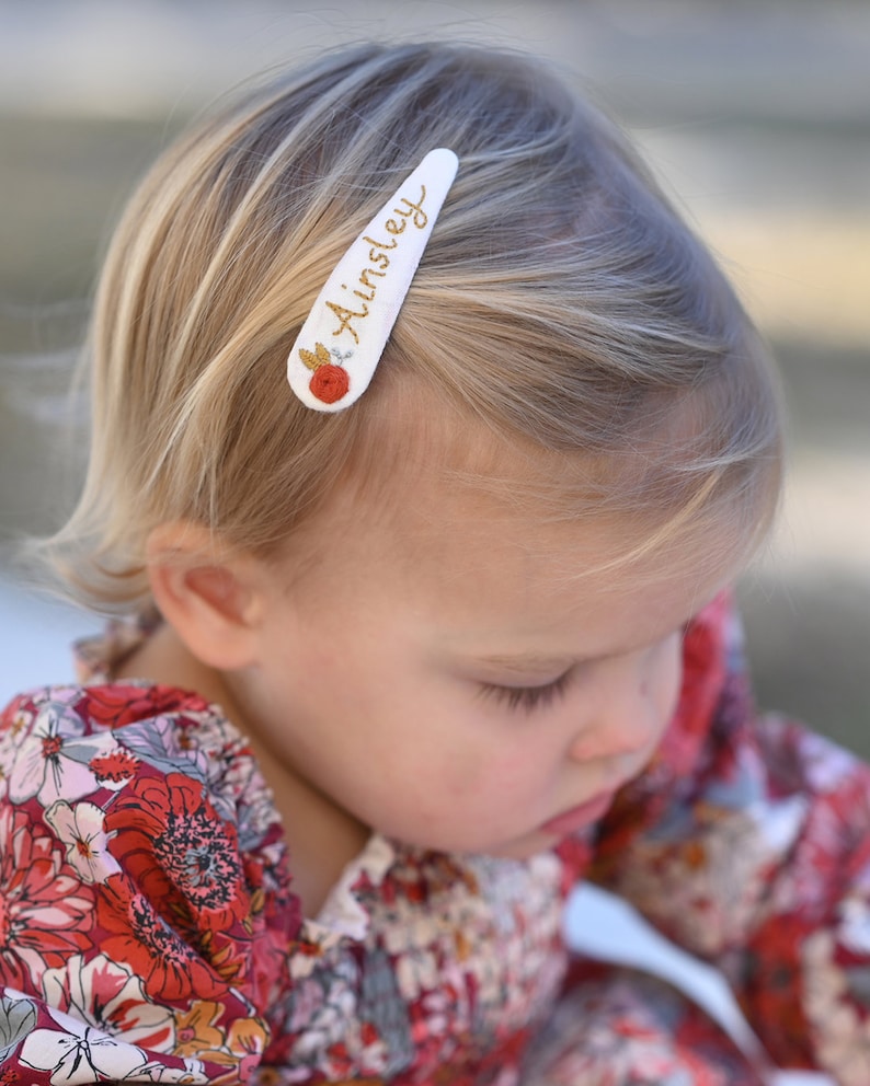 Pu&ograve; includere: Un fermaglio per capelli bianco con il nome "Ainsley" ricamato in filo dorato. Un piccolo fiore rosso con dettagli dorati &egrave; ricamato sul fermaglio.