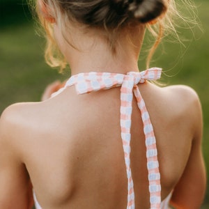 K&ouml;nnte beinhalten: Ein junges M&auml;dchen tr&auml;gt ein rosa-wei&szlig; kariertes Halfterkleid mit einer Schleife am Nacken.