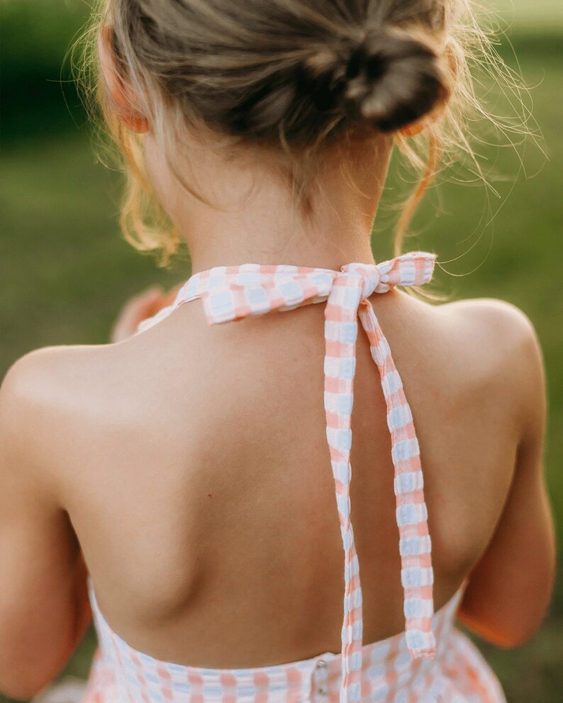 K&ouml;nnte beinhalten: Ein junges M&auml;dchen tr&auml;gt ein rosa-wei&szlig; kariertes Halfterkleid mit einer Schleife am Nacken.