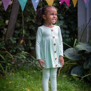 以下が含まれることがあります： 茶色のボタンが付いた薄い緑色の長袖シャツとそれに合わせたレギンスを着用した若い女の子。彼女は、カラフルな旗が頭上に吊るされた草地のエリアに立っている。