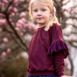 May include: A young child with blonde hair and blue eyes wearing a burgundy long-sleeved top with a velvet ruffle detail on the sleeve. The child is also wearing a blue skirt. The background features a blurred view of a tree with pink blossoms.