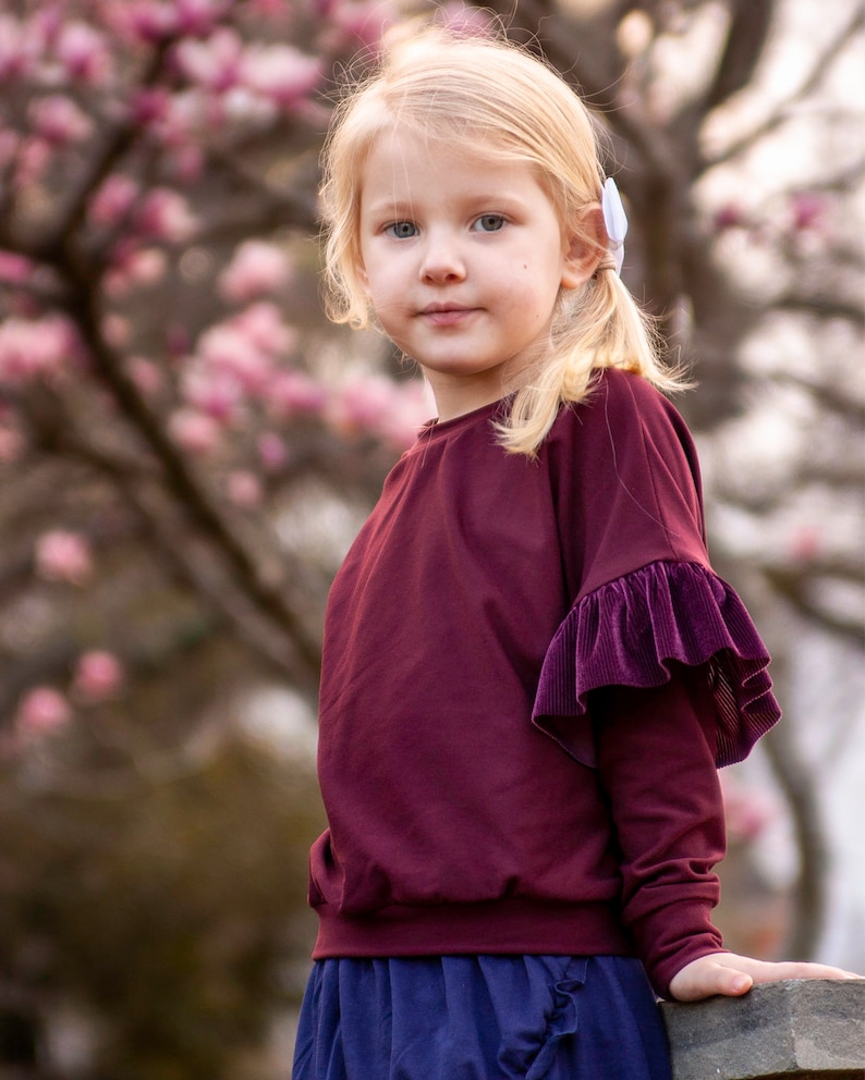 May include: A young child with blonde hair and blue eyes wearing a burgundy long-sleeved top with a velvet ruffle detail on the sleeve. The child is also wearing a blue skirt. The background features a blurred view of a tree with pink blossoms.