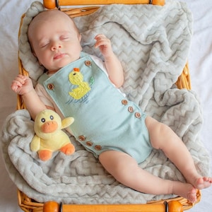 May include: A baby wearing a light blue romper with a yellow duck embroidered on the front, sleeping in a wicker basket lined with a gray chevron patterned blanket. A yellow duck plush toy is next to the baby.