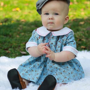May include: A baby wearing a blue and brown patterned dress with a white collar and pink stitching. The baby is wearing a gray newsboy cap and black leather shoes. The baby is sitting on a white fluffy surface.