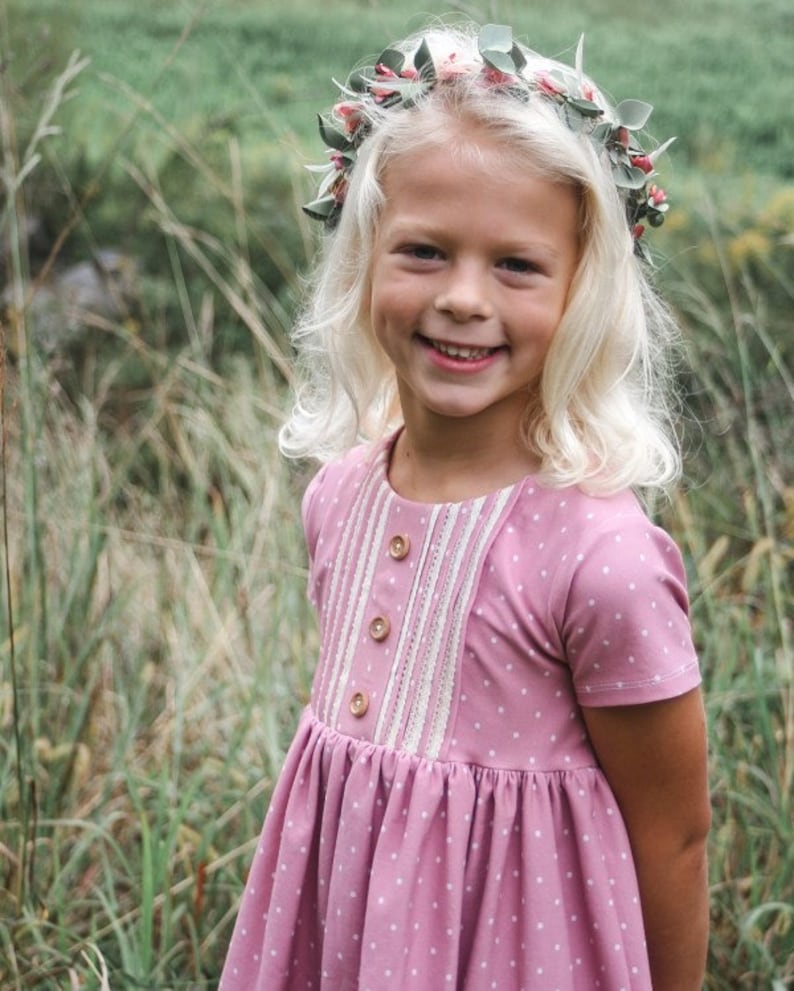 May include: A young girl wearing a pink polka dot dress with a white lace trim and wooden buttons. She is wearing a flower crown with green leaves and pink flowers.