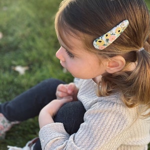 Pu&ograve; includere: Una bambina con i capelli castani indossa un maglione grigio chiaro e jeans blu scuro. Ha una molletta per capelli con motivo floreale nei capelli.