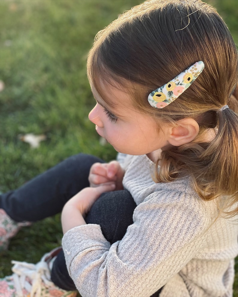 Pu&ograve; includere: Una bambina con i capelli castani indossa un maglione grigio chiaro e jeans blu scuro. Ha una molletta per capelli con motivo floreale nei capelli.