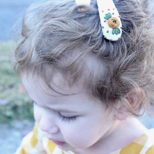Pu&ograve; includere: Una bambina con i capelli biondi ricci che indossa una camicia a quadri gialla e bianca con un fermaglio per capelli con un fiore bianco ricamato.