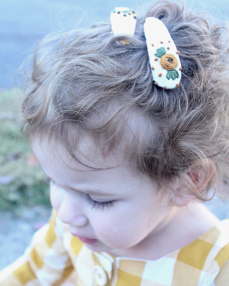 Pu&ograve; includere: Una bambina con i capelli biondi ricci che indossa una camicia a quadri gialla e bianca con un fermaglio per capelli con un fiore bianco ricamato.