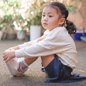 May include: A young child wearing a cream-colored long-sleeved shirt, navy blue shorts, and white sneakers with a pink swoosh logo. The child is sitting on a gray surface, with a blurred background of greenery.