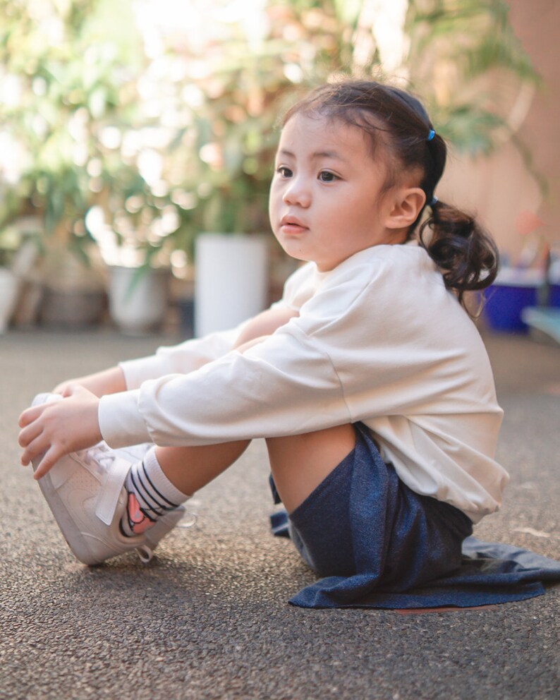 May include: A young child wearing a cream-colored long-sleeved shirt, navy blue shorts, and white sneakers with a pink swoosh logo. The child is sitting on a gray surface, with a blurred background of greenery.