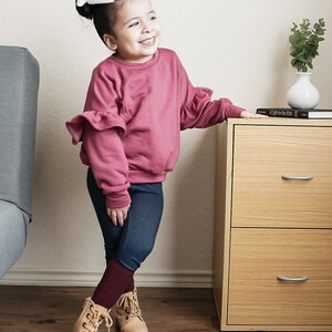 May include: A young child wearing a mauve-colored sweatshirt with ruffled sleeves, dark blue leggings, and tan lace-up boots. A large white bow adorns the child's hair. The child is smiling and leaning against a wooden cabinet.