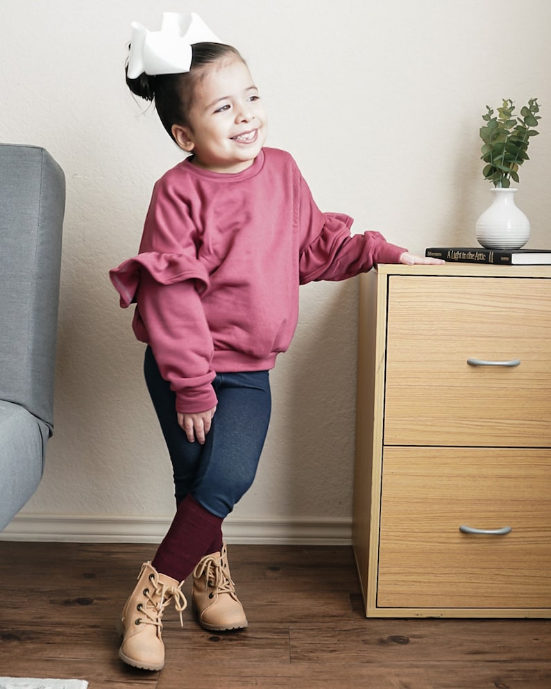 May include: A young child wearing a mauve-colored sweatshirt with ruffled sleeves, dark blue leggings, and tan lace-up boots. A large white bow adorns the child's hair. The child is smiling and leaning against a wooden cabinet.
